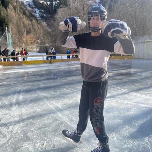 Ein Schüler mit Helm, Handschuhe und Eislaufschuhe auf dem Eis. Im Hintergrund die Schüler am Zaun.