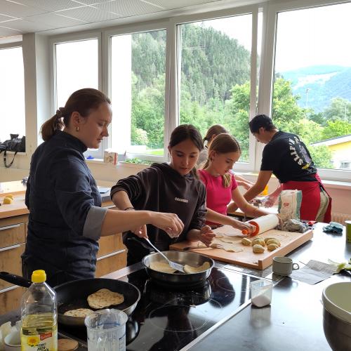 Die Lehrerin zeigt einer Mittelschülerin, wie man Pita in der Pfanna macht, im Hintergrund Arbeiten an der Nudelmaschine