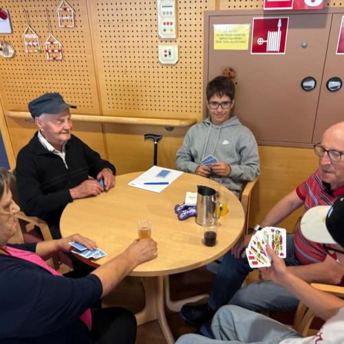 Zwei Schüler spielen an einem runden Tisch mit drei Bewohner des Pflegeheims Ried Karten.